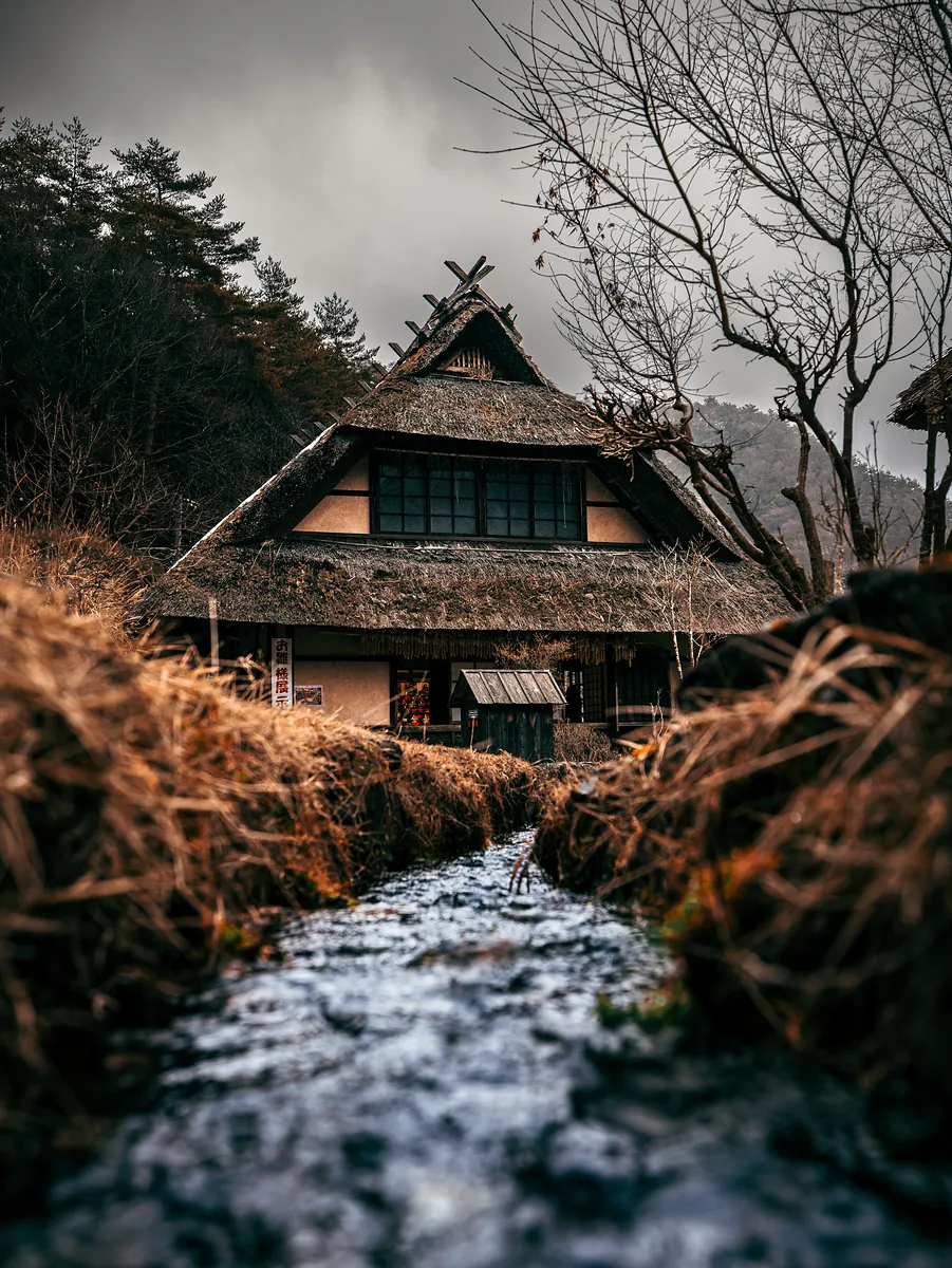 A thatched-roof farmhouse behind a narrow stream lined with dry grass, under a grey sky.