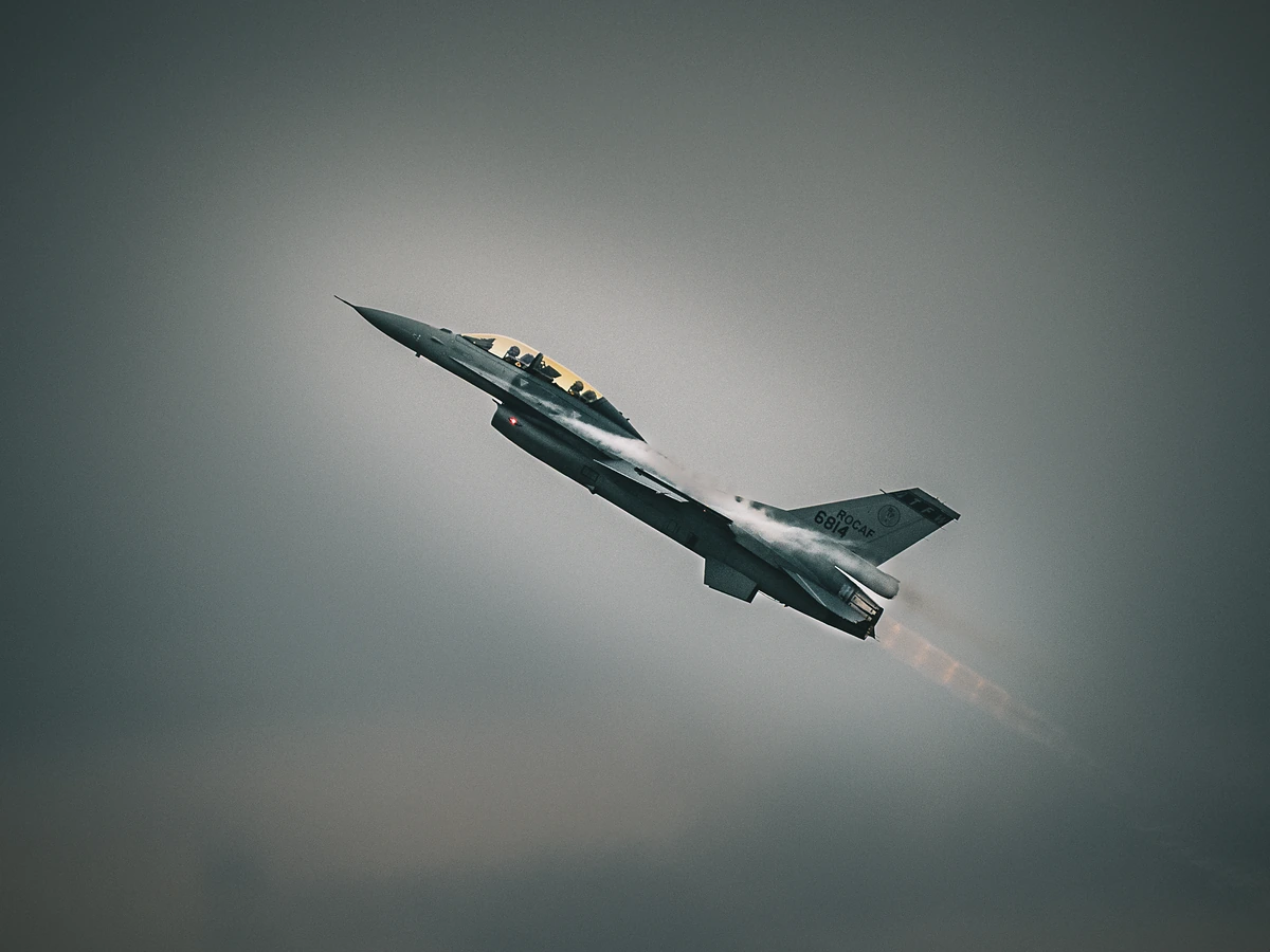 A two-seat F-16 climbs steeply in grey sky, trailing vapor and afterburner glow.