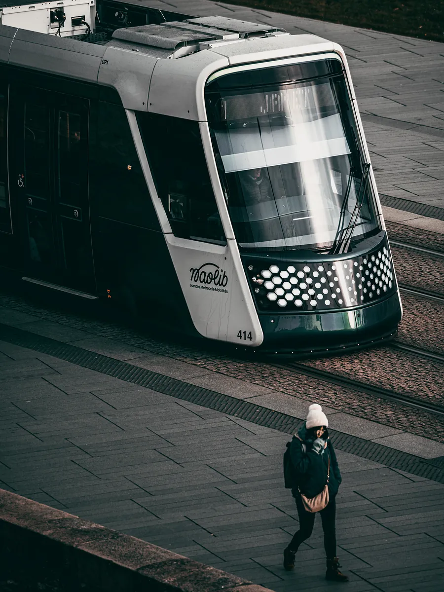 A dark green Nantes tram glides past as a woman in a white knit hat walks across the square.