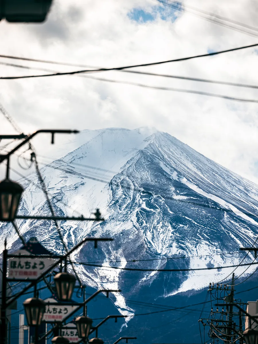 Mount Fuji's snowy flank seen through overhead wires and street lamps, with Honcho street signs below.
