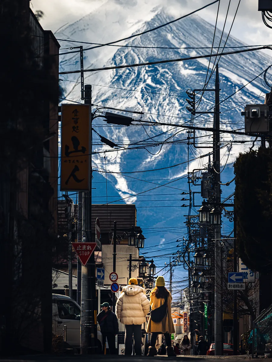 People in winter coats stand in a street strung with signs and power lines while Mount Fuji fills the sky behind them.