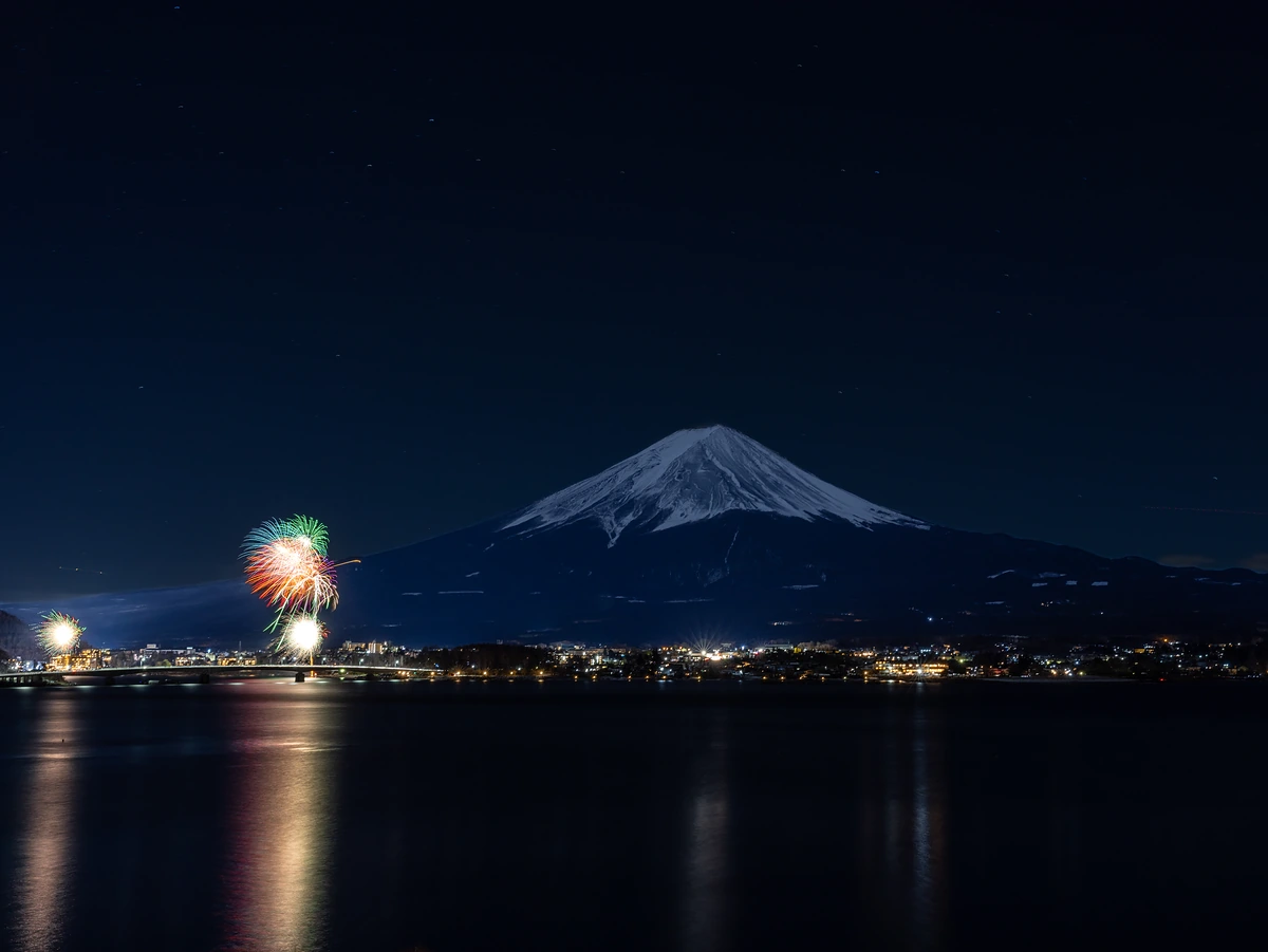Mount Fuji at night with fireworks bursting over Lake Kawaguchi and town lights reflected in the water.