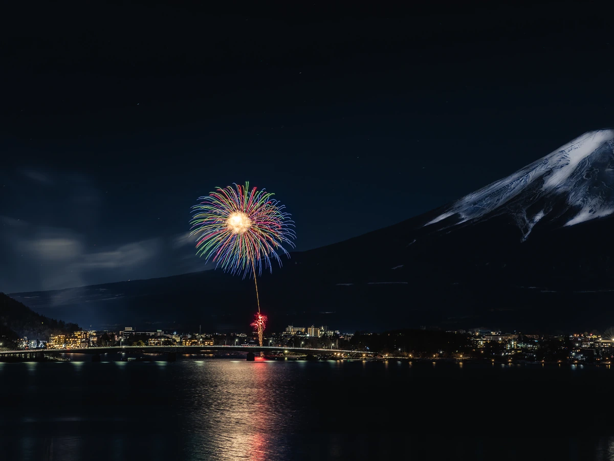 A single firework bursts over Lake Kawaguchi at night, reflected in the water, with Fuji's snowy slope at right.