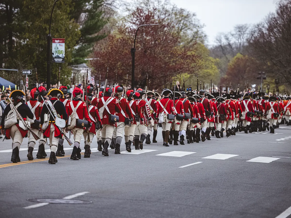 A long column of redcoat reenactors carrying muskets marches down a street lined with spectators.