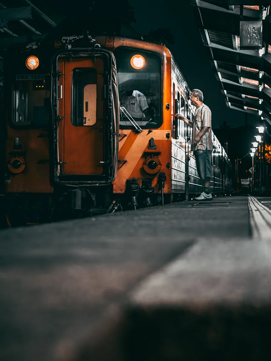 An orange diesel railcar with glowing headlights at a night platform, a man in a striped shirt at its door.