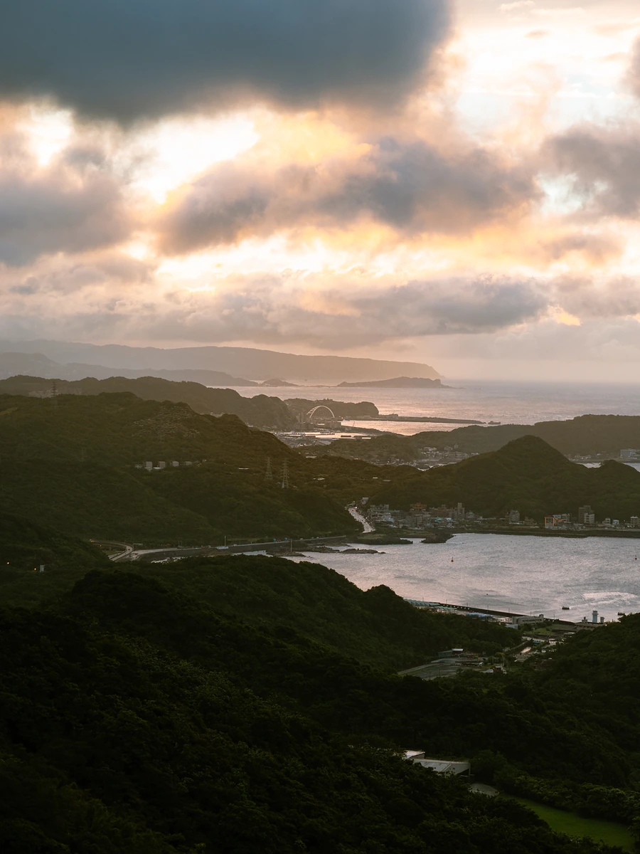 Green hills stepping down to a bay and the sea at sunset, under glowing broken cloud.