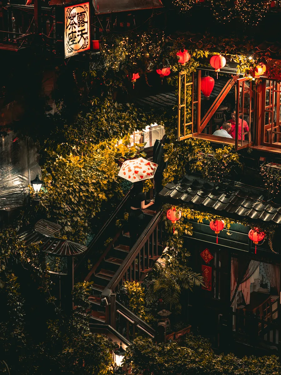 Night view of a teahouse's stairs and balconies lit by red lanterns, a woman with a polka-dot umbrella climbing the steps.