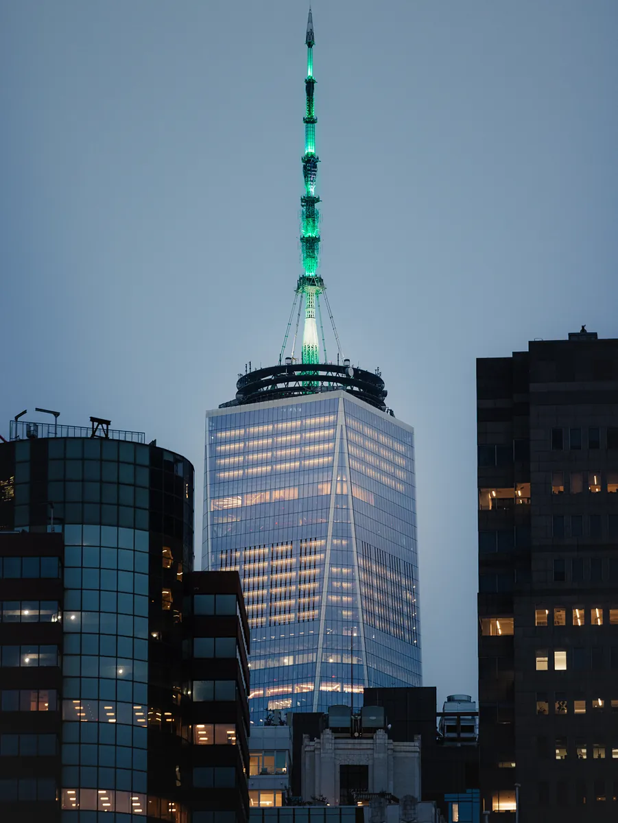 One World Trade Center at blue hour, its spire lit green above darker office towers.