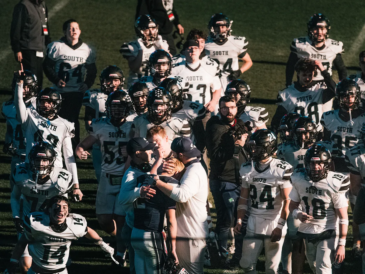 A football team in white jerseys spills across the field in low sun while coaches embrace a player.