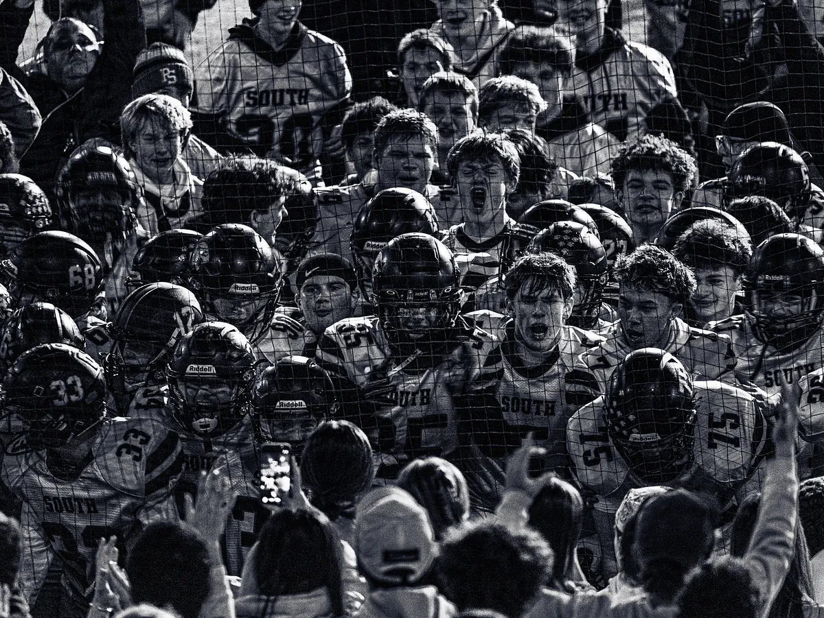 Black-and-white view through a net of a football team celebrating with fans, players shouting.