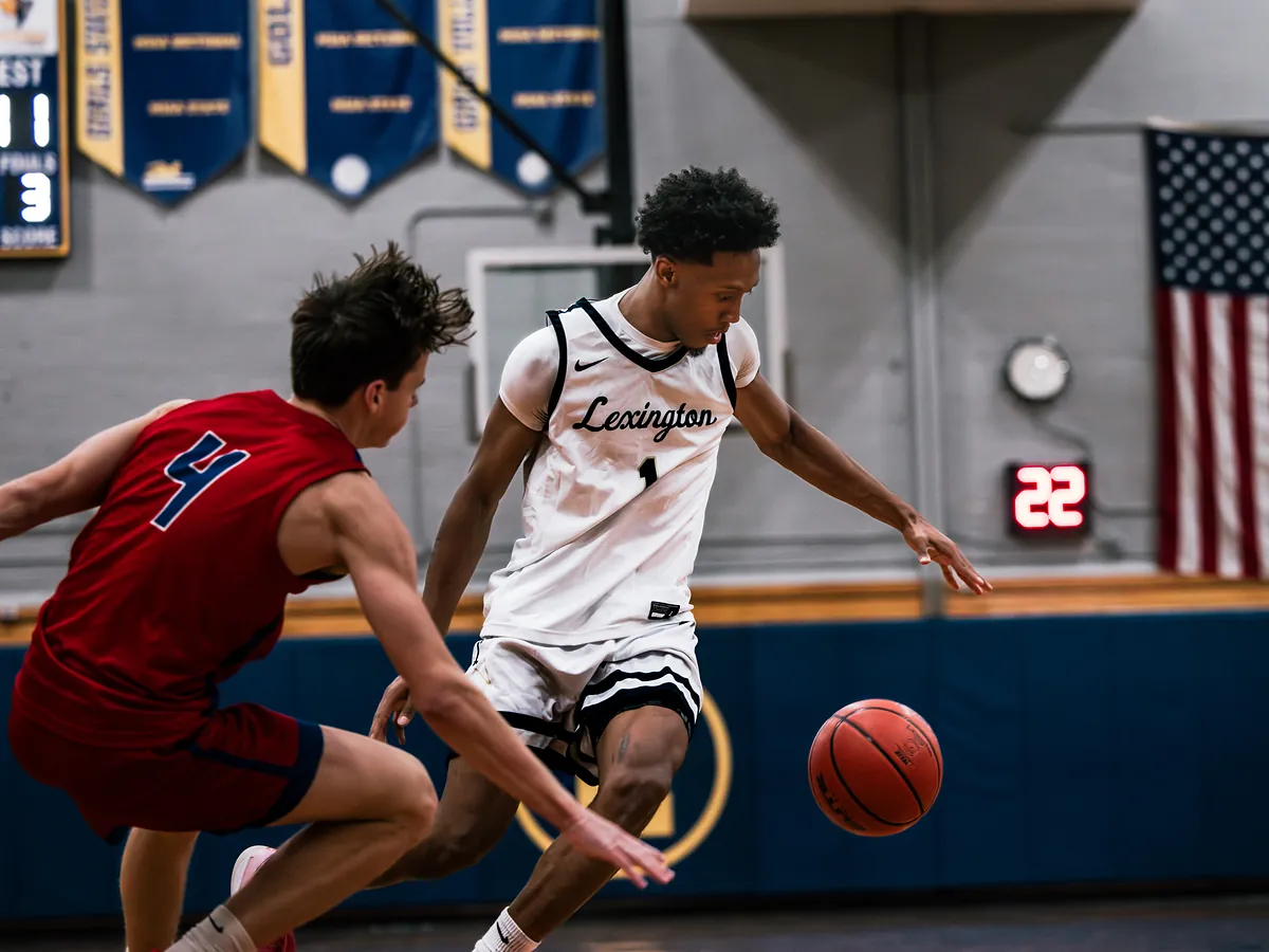 A basketball player in white drives past a defender in red, the ball bouncing low.
