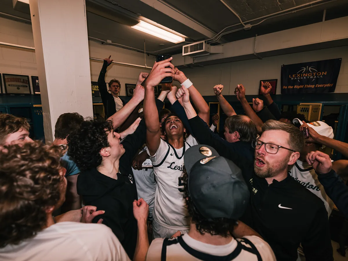 A crowded locker room of players and coaches cheering with fists raised, phones held high.
