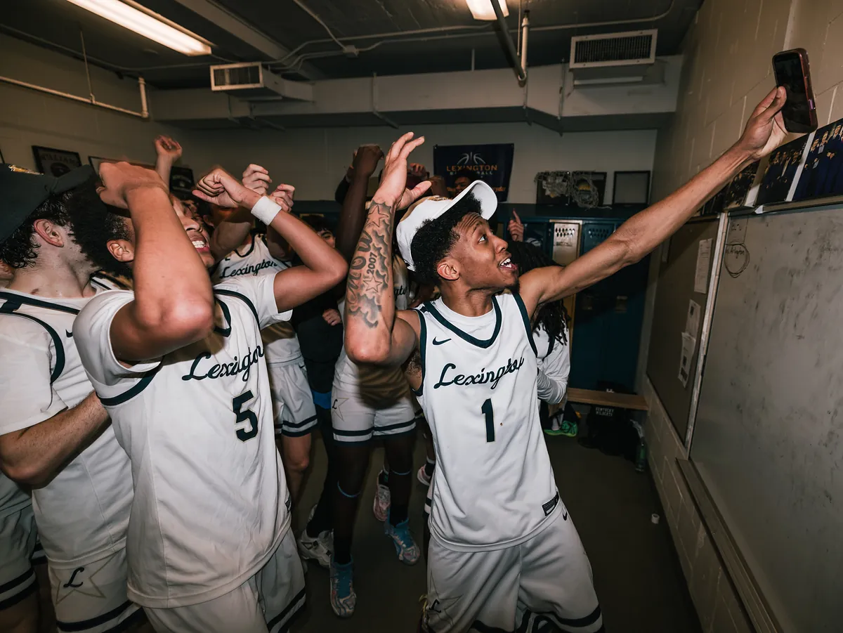 Players dance and film on their phones in a locker room, one stretching his arm out for a selfie.