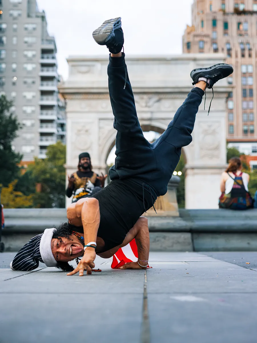 A breakdancer holds a one-handed freeze on the pavement, grinning at the camera, with the arch behind him.