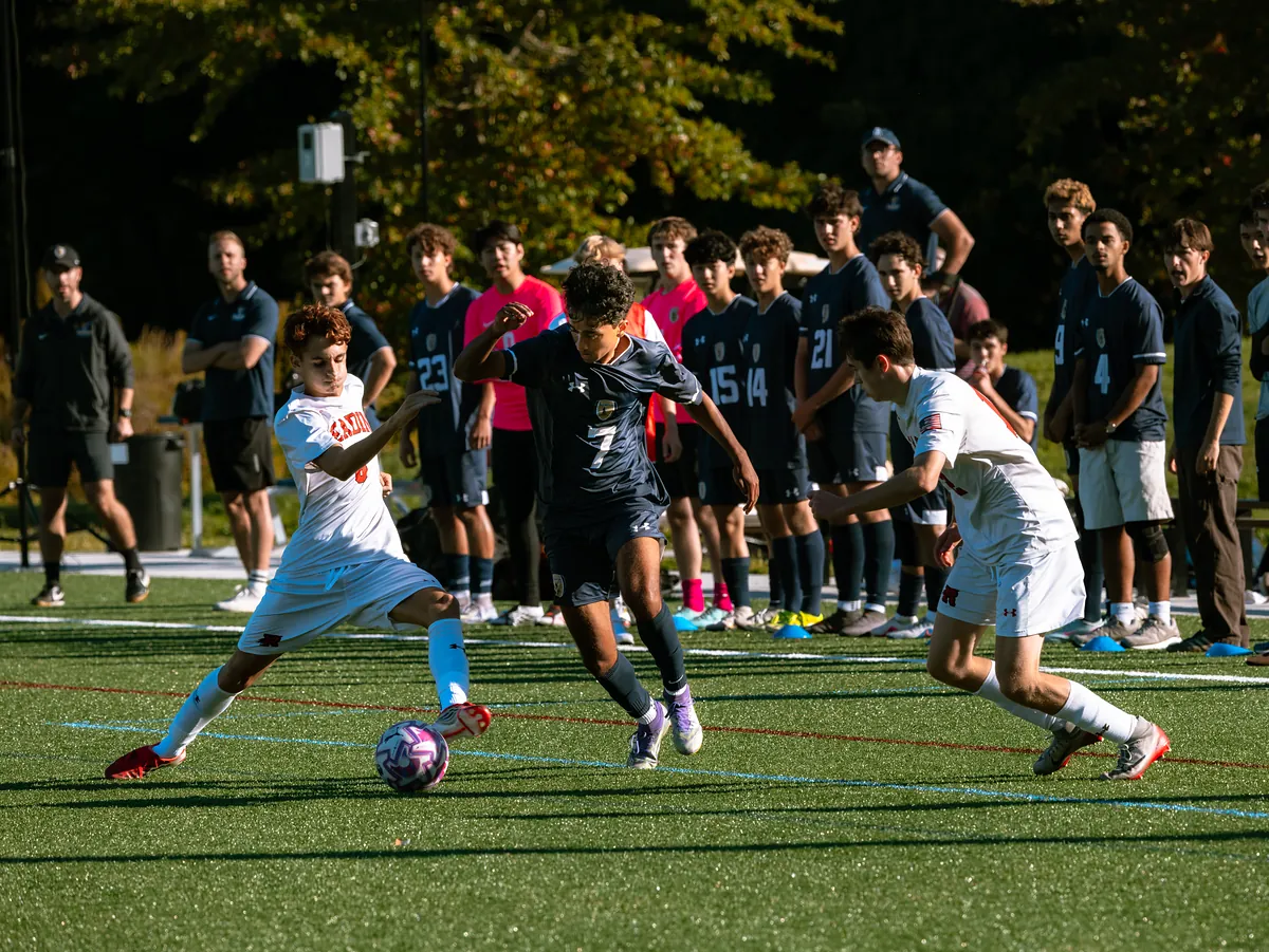 A soccer player in navy dribbles between two defenders in white as teammates watch from the sideline.